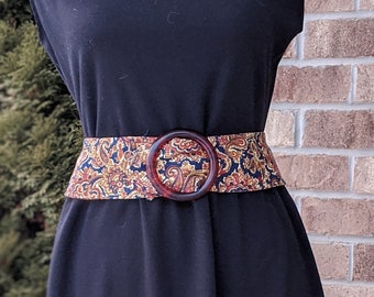 Pode incluir: Um vestido preto com um cinto estampado. O cinto apresenta um design paisley em tons de ouro, vermelho e azul, e tem uma fivela grande e redonda castanha. O vestido é sem mangas e tem uma bainha ao nível do joelho.