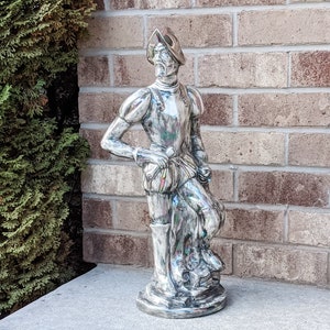 May include: A silver-colored ceramic statue of a man in a conquistador costume. The statue is standing on a base and has a detailed design.