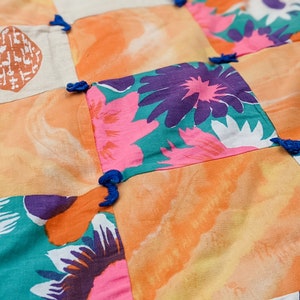 Puede incluir: Una colcha de retazos con cuadrados de diferentes patrones florales en tonos de rosa, azul, verde y naranja. La colcha está atada con borlas azules.