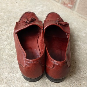 Pode incluir: Um par de mocassins de couro marrom com franjas e sola marrom escura. Os mocassins t&ecirc;m bico fino e um detalhe de franjas na parte superior.