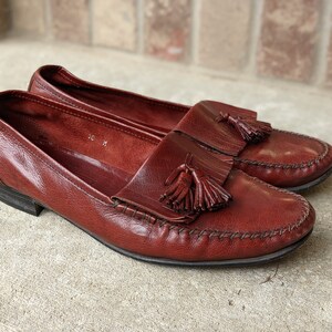 Pode incluir: Um par de mocassins de couro marrom com franjas e um detalhe costurado ao redor da ponta. Os mocassins t&ecirc;m um pequeno salto e uma franja em cada sapato.