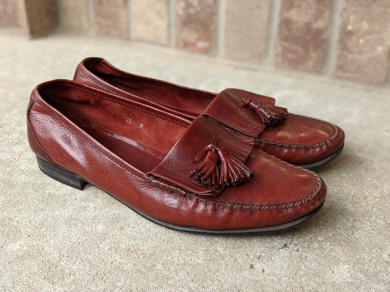 Pode incluir: Um par de mocassins de couro marrom com franjas e um detalhe costurado ao redor da ponta. Os mocassins t&ecirc;m um pequeno salto e uma franja em cada sapato.