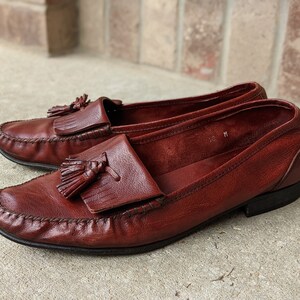 Pode incluir: Um par de mocassins de couro marrom com franjas e bico fino. Os mocassins t&ecirc;m um ponto decorativo ao redor da borda da sola e uma franja na parte superior do peito do p&eacute;.