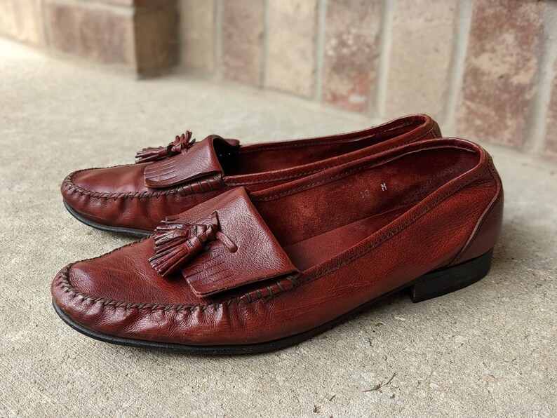 Pode incluir: Um par de mocassins de couro marrom com franjas e bico fino. Os mocassins t&ecirc;m um ponto decorativo ao redor da borda da sola e uma franja na parte superior do peito do p&eacute;.