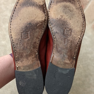 Pode incluir: Close-up das solas de um par de sapatos de couro marrom. As solas s&atilde;o de couro marrom e t&ecirc;m uma banda de rodagem de borracha preta. Os sapatos s&atilde;o etiquetados como "HAND MADE IN ITALY" e "VIBRAM".