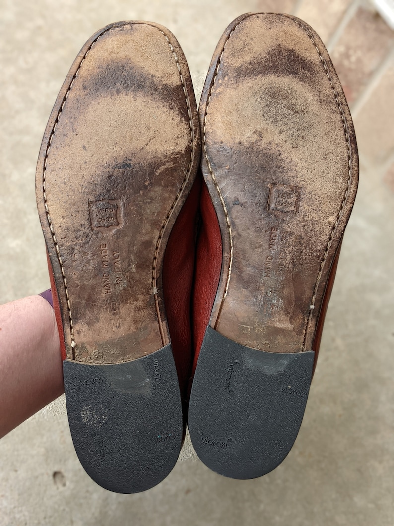 Pode incluir: Close-up das solas de um par de sapatos de couro marrom. As solas s&atilde;o de couro marrom e t&ecirc;m uma banda de rodagem de borracha preta. Os sapatos s&atilde;o etiquetados como "HAND MADE IN ITALY" e "VIBRAM".
