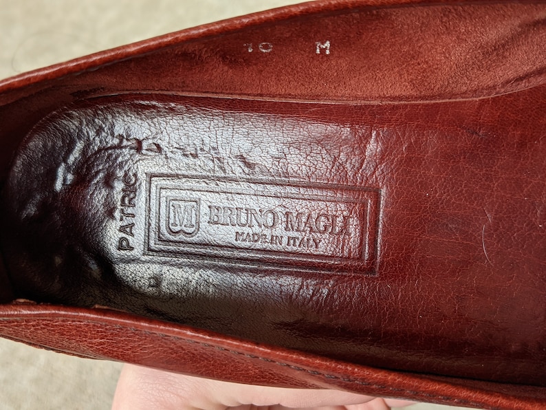 Pode incluir: Close-up do interior de um sapato de couro marrom com o texto "Bruno Magli Made in Italy" gravado na palmilha.