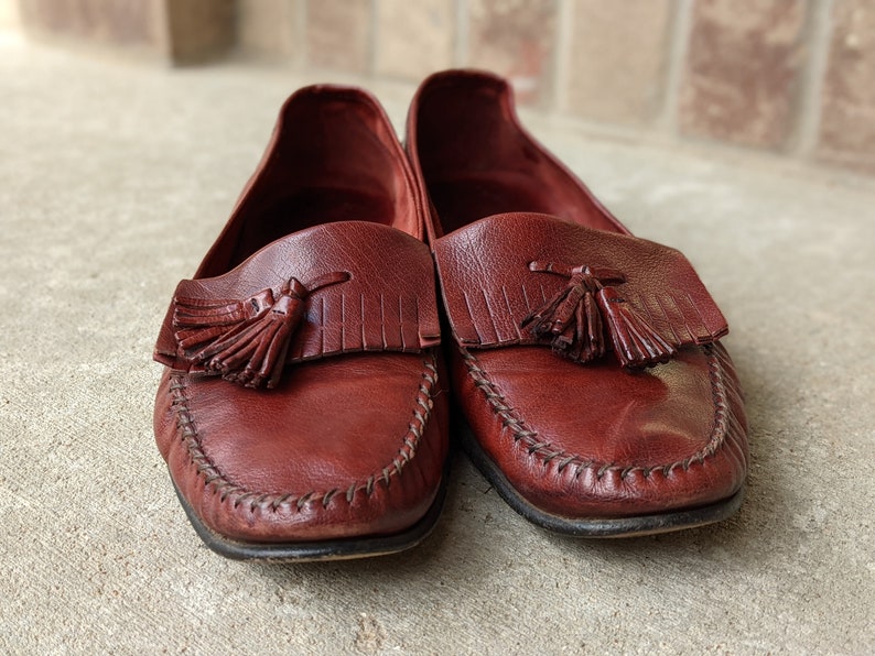 Pode incluir: Um par de mocassins de couro castanho com borlas. Os mocassins t&ecirc;m uma costura em volta da borda e uma ponta pontiaguda.