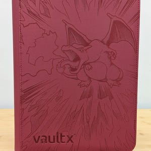 May include: A dark red zippered trading card binder featuring an embossed graphic of a dragon-like creature breathing fire on the front cover. The brand name vaultx is embossed in the bottom left corner of the textured, rectangular protective case.