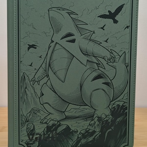 May include: A sage green zippered binder featuring a detailed black line art illustration of a large, bipedal, armored creature standing on a rocky cliffside with birds flying in the sky. The word Vault X is embossed in the bottom left corner.