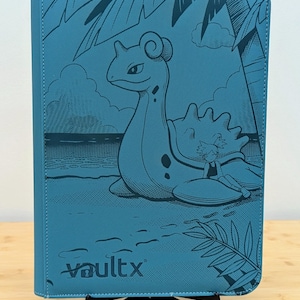 May include: A teal blue trading card binder featuring a dark blue line art illustration of a Lapras creature and a character on a beach with palm trees. The front cover displays the VaultX logo in the bottom left corner with a textured finish.