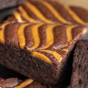 May include: Close-up of a stack of chocolate brownies with a swirl of peanut butter on top. The brownies are dark brown and the peanut butter is a light brown.