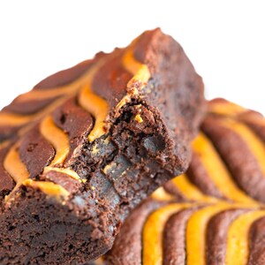 May include: A close-up of a chocolate brownie with a swirl of caramel on top. The brownie is broken in half, revealing the soft, fudgy inside.