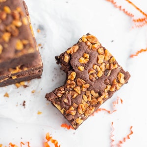 May include: A close-up of a chocolate brownie with pecans, a bite taken out of it. The brownie is on a white surface with orange curling ribbon.