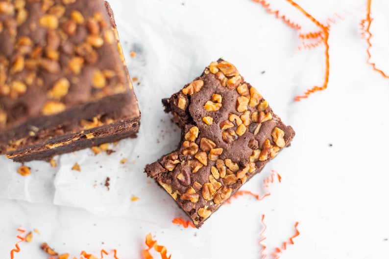 May include: A close-up of a chocolate brownie with pecans, a bite taken out of it. The brownie is on a white surface with orange curling ribbon.