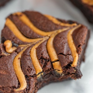 May include: A close-up of a chocolate brownie with peanut butter swirls. The brownie has a bite taken out of it, revealing the soft, gooey inside.