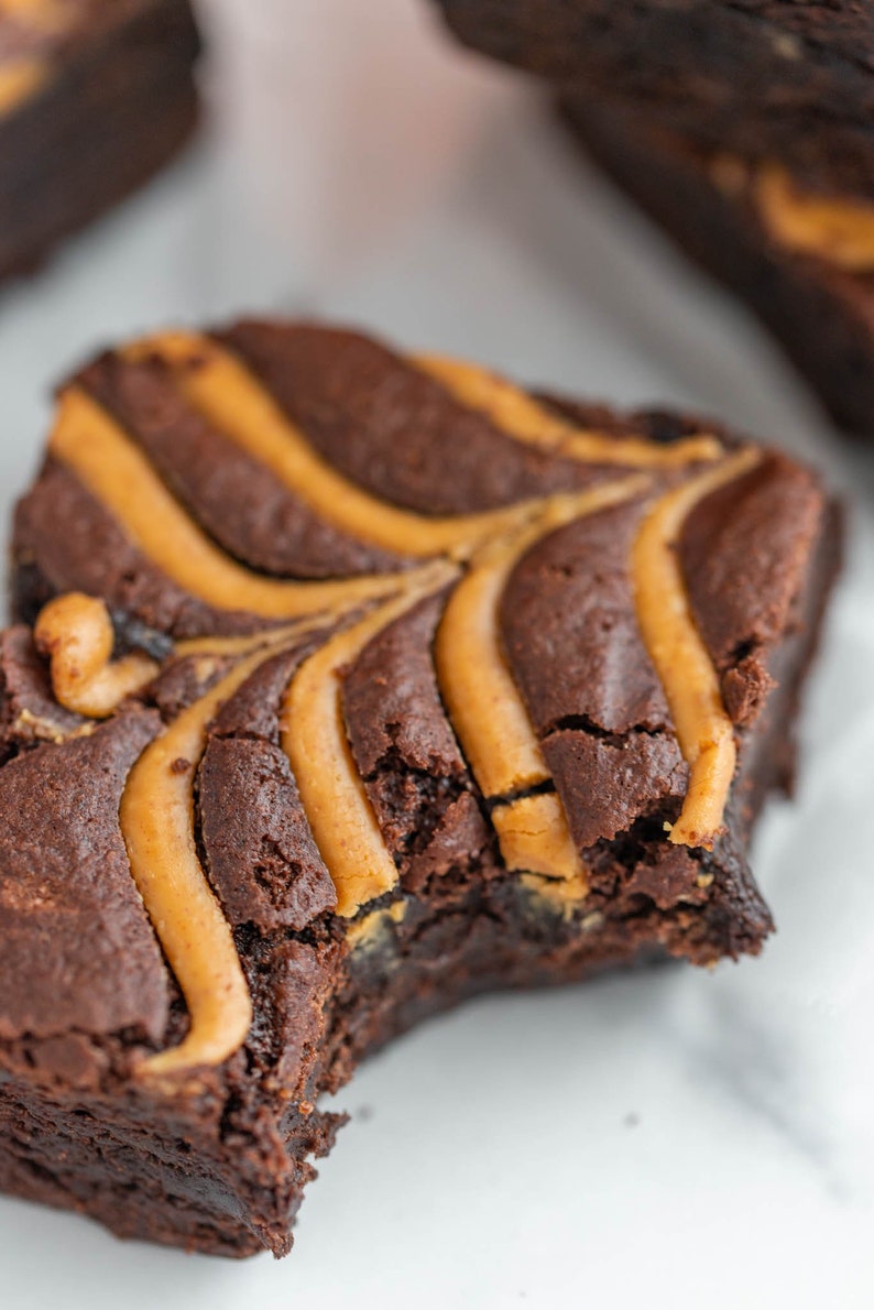 May include: A close-up of a chocolate brownie with peanut butter swirls. The brownie has a bite taken out of it, revealing the soft, gooey inside.