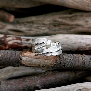 May include: A silver ring with a feather design. The ring has detailed feather textures and wraps around the finger. The ring is displayed on a piece of wood, with a blurred wooden background.