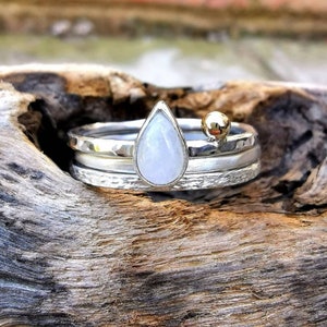 May include: A silver ring set with three bands, featuring a teardrop-shaped moonstone and a small gold bead.