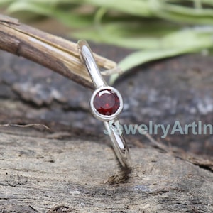 Op de afbeelding: Een zilveren ring met een kleine, ronde, rode edelsteen in het midden.
