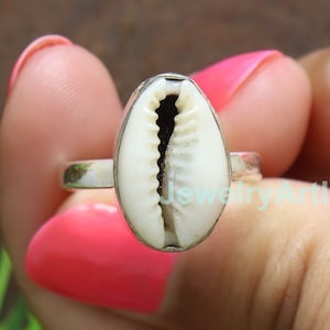 May include: A silver ring featuring a white and beige cowrie shell. The shell is oval-shaped and set in a silver band. The ring is held by a person with pink nail polish.