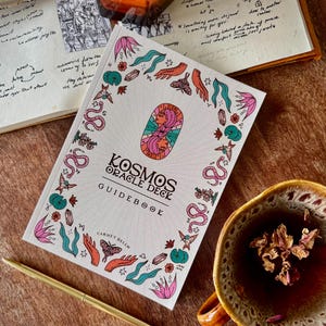 Puede incluir: Una guía blanca de la baraja del Oráculo Kosmos con ilustraciones y texto coloridos. La portada presenta un diseño central de dos caras y está rodeada de imágenes botánicas y celestiales. También se ven una vela encendida, un bolígrafo y una taza de té.