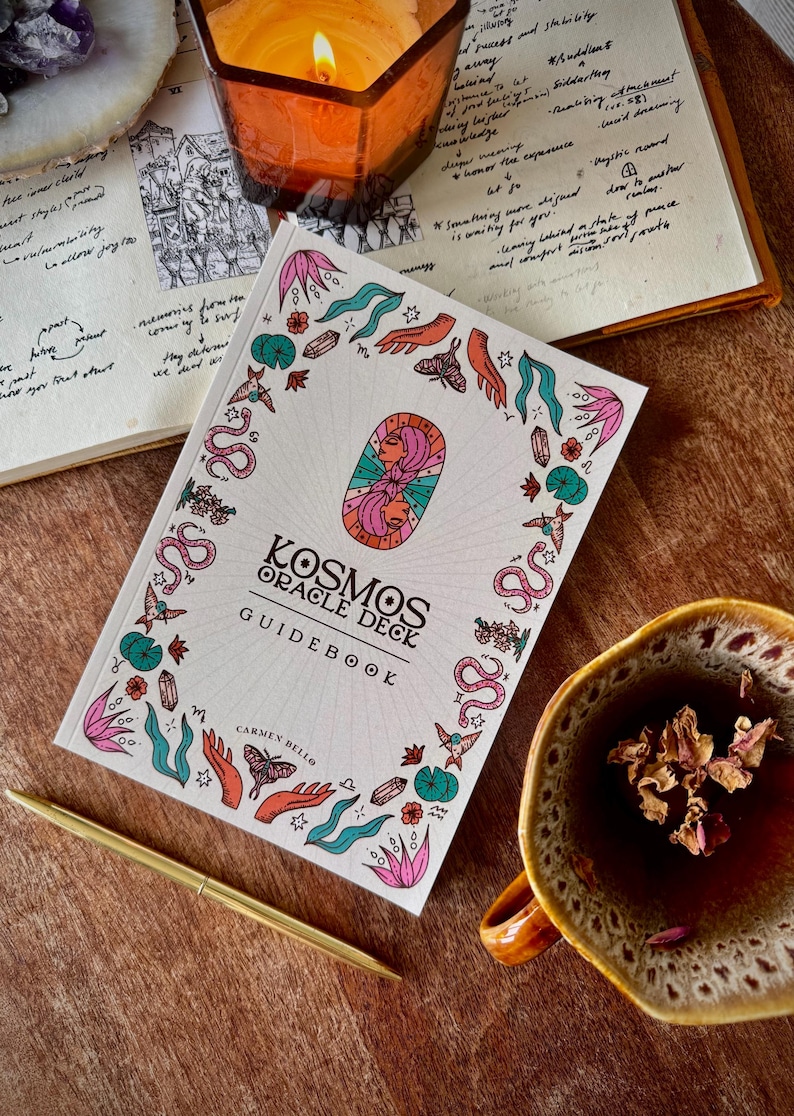 May include: A white Kosmos Oracle Deck guidebook with colorful illustrations and text. The cover features a central design of two faces and is surrounded by botanical and celestial imagery. A lit candle, pen, and cup of tea are also visible.