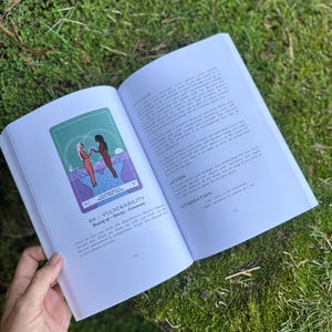May include: An open book with a green and purple illustration of two figures holding hands. The text on the page reads "VULNERABILITY" and "Showing up - Honesty - Communion". The book is held open on a mossy surface.