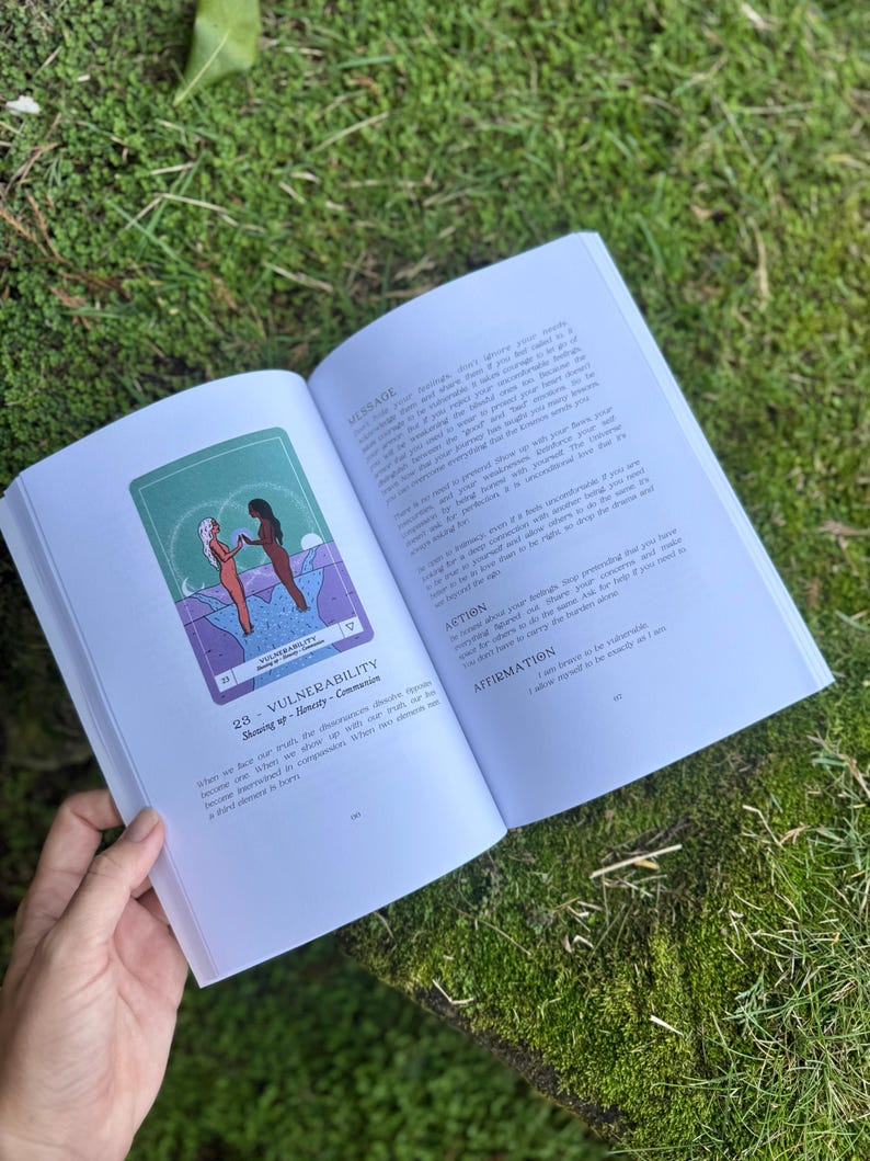 May include: An open book with a green and purple illustration of two figures holding hands. The text on the page reads "VULNERABILITY" and "Showing up - Honesty - Communion". The book is held open on a mossy surface.
