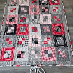 May include: A rectangular patchwork quilt featuring a grid of squares in red, black, white, and gray. The fabric patterns include gingham, polka dots, and stripes. The quilt is bordered by a solid gray fabric and includes a white braided tie.
