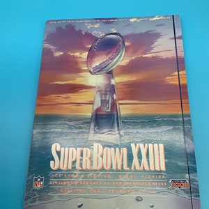 May include: A program for Super Bowl XXIII, featuring a colorful illustration of the Vince Lombardi Trophy against a sunset backdrop. The text reads "Super Bowl XXIII" and "Joe Robbie Stadium, Miami, Florida" with the teams playing, "Cincinnati Bengals vs. San Francisco 49ers".