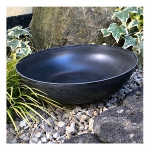 Könnte beinhalten: Eine große, schwarze, flache Schale mit strukturierter Außenseite und glatter, reflektierender Innenseite. Die Schale hat einen dünnen, silberfarbenen Rand. Sie steht zwischen Felsen, Grün und Blättern, was auf eine Umgebung im Freien hindeutet.