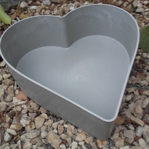 Puede incluir: Una jardinera gris en forma de corazón hecha de un material duradero. La jardinera tiene un interior liso y un exterior ligeramente texturizado. Los bordes son limpios y definidos, con un borde blanco. La jardinera está diseñada para uso en exteriores.