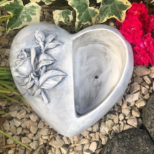 Könnte beinhalten: Ein grauer herzförmiger Pflanzer mit einem Rosenmuster auf der einen Seite und einem Abzugsloch auf der anderen. Der Pflanzer ist aus Beton und hat eine rustikale Oberfläche.