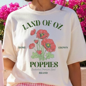 May include: Ivory t-shirt with a "Land of Oz Poppies" graphic, paired with pink gingham shorts and a pink baseball cap. A pink Stanley tumbler is held in the hand. The background features vibrant pink flowers and a clear blue sky.