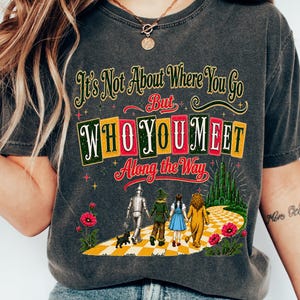 May include: A charcoal gray short-sleeve t-shirt featuring a graphic print of characters walking on a yellow brick road. The text reads: It's Not About Where You Go But WHO YOU MEET Along the Way. The design includes red poppies and a green castle.