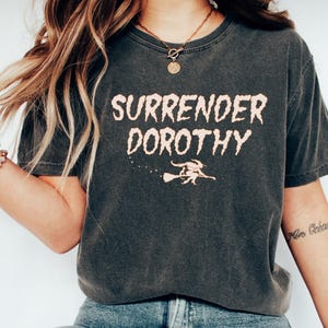 May include: Dark gray t-shirt with the text "SURRENDER DOROTHY" in a distressed, light orange font. Below the text is a graphic of a witch on a broom. The shirt is paired with blue jeans and white shoes.