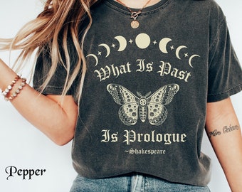 May include: A charcoal gray short-sleeve t-shirt featuring a cream-colored graphic of moon phases, a moth, and the text "What Is Past Is Prologue ~Shakespeare". The shirt has a faded, vintage-style wash and is paired with blue denim jeans.