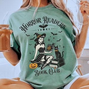 May include: A light green t-shirt with a black graphic of a witch holding a book with a black cat sitting on a stack of books. The text on the t-shirt reads 'Horror Readers Book Club'.