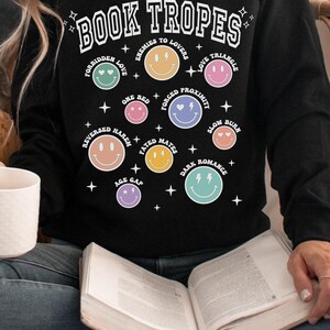 May include: Black sweatshirt with the text "BOOK TROPES" and various smiley face graphics representing different romance novel tropes. The sweatshirt includes phrases like "Enemies to Lovers" and "Love Triangle".