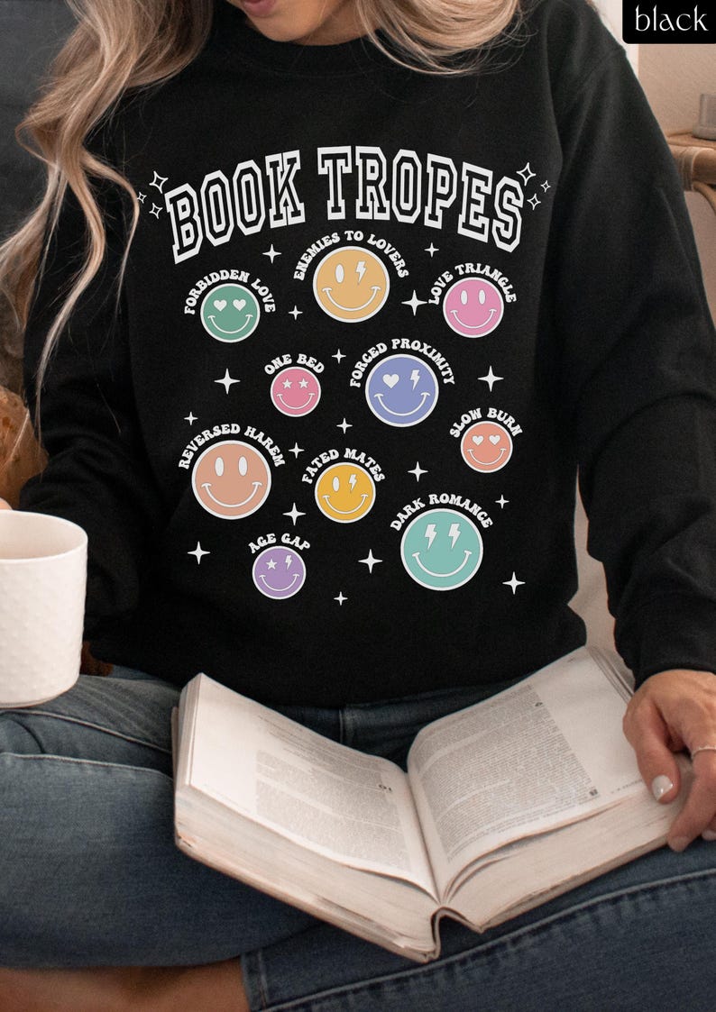 May include: Black sweatshirt with the text "BOOK TROPES" and various smiley face graphics representing different romance novel tropes. The sweatshirt includes phrases like "Enemies to Lovers" and "Love Triangle".