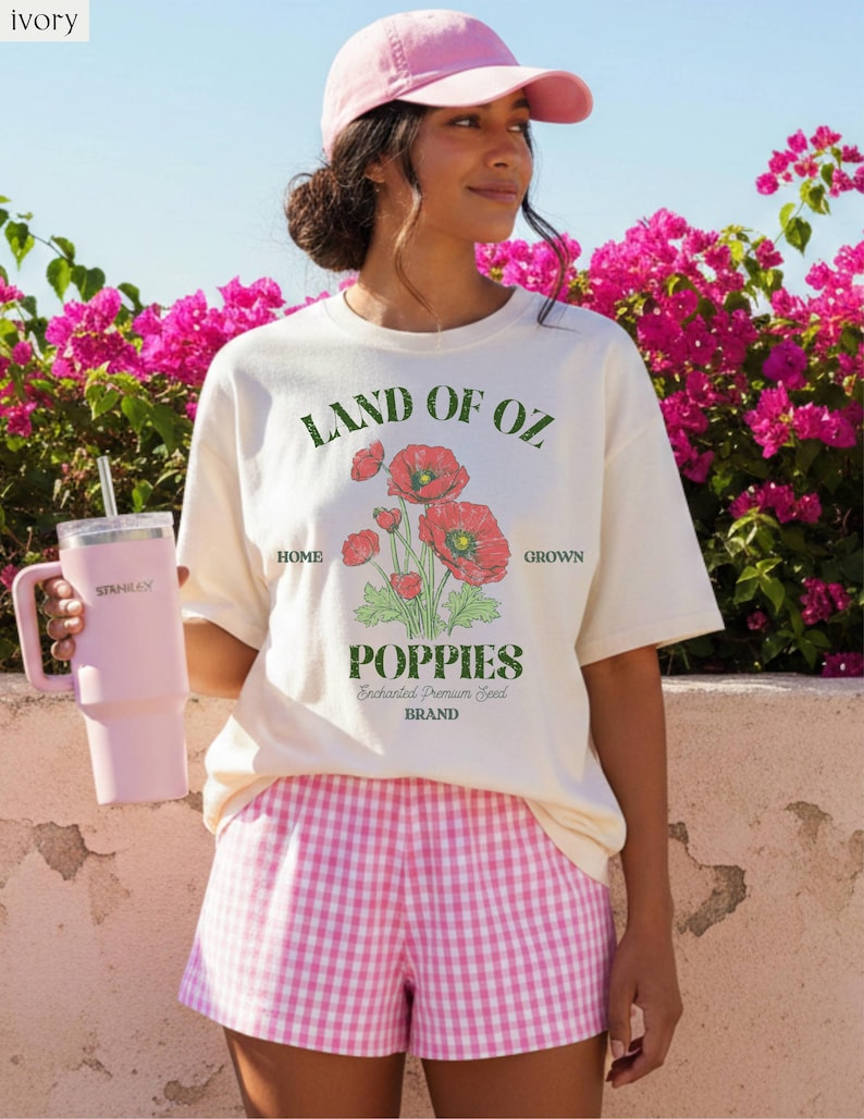 May include: Ivory t-shirt with a "Land of Oz Poppies" graphic, paired with pink gingham shorts and a pink baseball cap. A pink Stanley tumbler is held in the hand. The background features vibrant pink flowers and a clear blue sky.