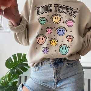 May include: Beige sweatshirt with the text "BOOK TROPES" and various smiley face graphics representing book tropes like "Forbidden Love" and "Enemies to Lovers." The sweatshirt is paired with light-wash ripped jeans.