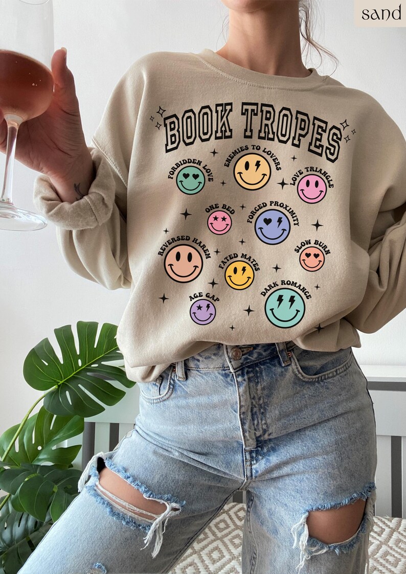 May include: Beige sweatshirt with the text "BOOK TROPES" and various smiley face graphics representing book tropes like "Forbidden Love" and "Enemies to Lovers." The sweatshirt is paired with light-wash ripped jeans.