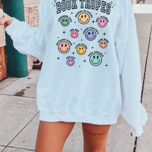 May include: White sweatshirt with the text "BOOK TROPES" in black, with various smiley face graphics and phrases like "Enemies to Lovers" and "Forbidden Love". The sweatshirt is a casual style.
