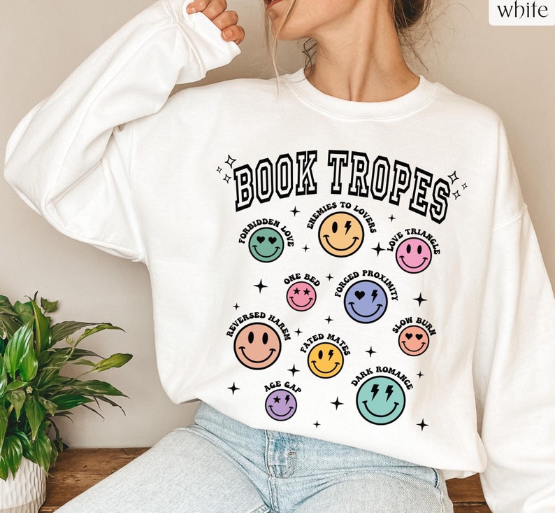 May include: White sweatshirt with the text "BOOK TROPES" in black, with various smiley faces representing book tropes like "Forbidden Love" and "Enemies to Lovers." The design includes stars and a retro aesthetic.