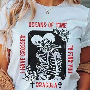 May include: White t-shirt with a black and white graphic of two skeletons embracing. The text "OCEANS OF TIME I HAVE CROSSED TO FIND YOU" is written in red above the graphic. The text "DRACULA" is written in red below the graphic.