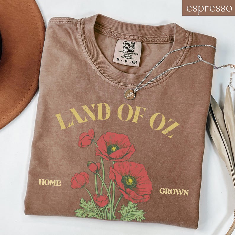 May include: Espresso-colored t-shirt with the text "LAND OF OZ" in yellow, and a floral graphic of red poppies. The shirt also has the words "HOME" and "GROWN". A silver necklace with a pendant is draped across the shirt.