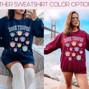 May include: Navy blue and maroon sweatshirts with the text "BOOK TROPES" and colorful smiley face graphics. The sweatshirts have a crew neck and long sleeves. The navy sweatshirt is paired with white knee-high socks.