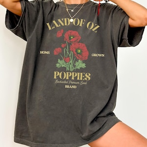 May include: Dark gray oversized t-shirt with a graphic featuring red poppies and the text "LAND OF OZ POPPIES." The design includes the words "HOME GROWN" and "Enchanted Premium Seed BRAND" in a vintage style.